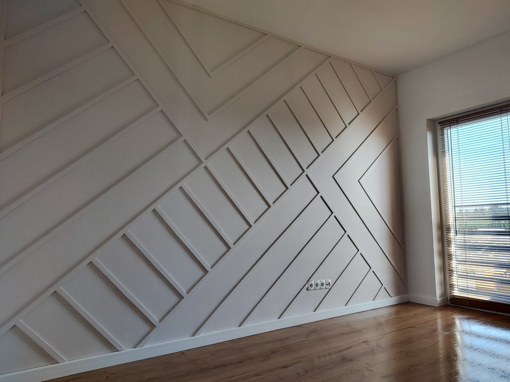 Ściana z dekoracyjnymi, geometrycznymi panelami MDF w kolorze jasnego beżu, z widocznymi gniazdkami elektrycznymi i fragmentem podłogi z paneli drewnopodobnych, oświetlona naturalnym światłem...