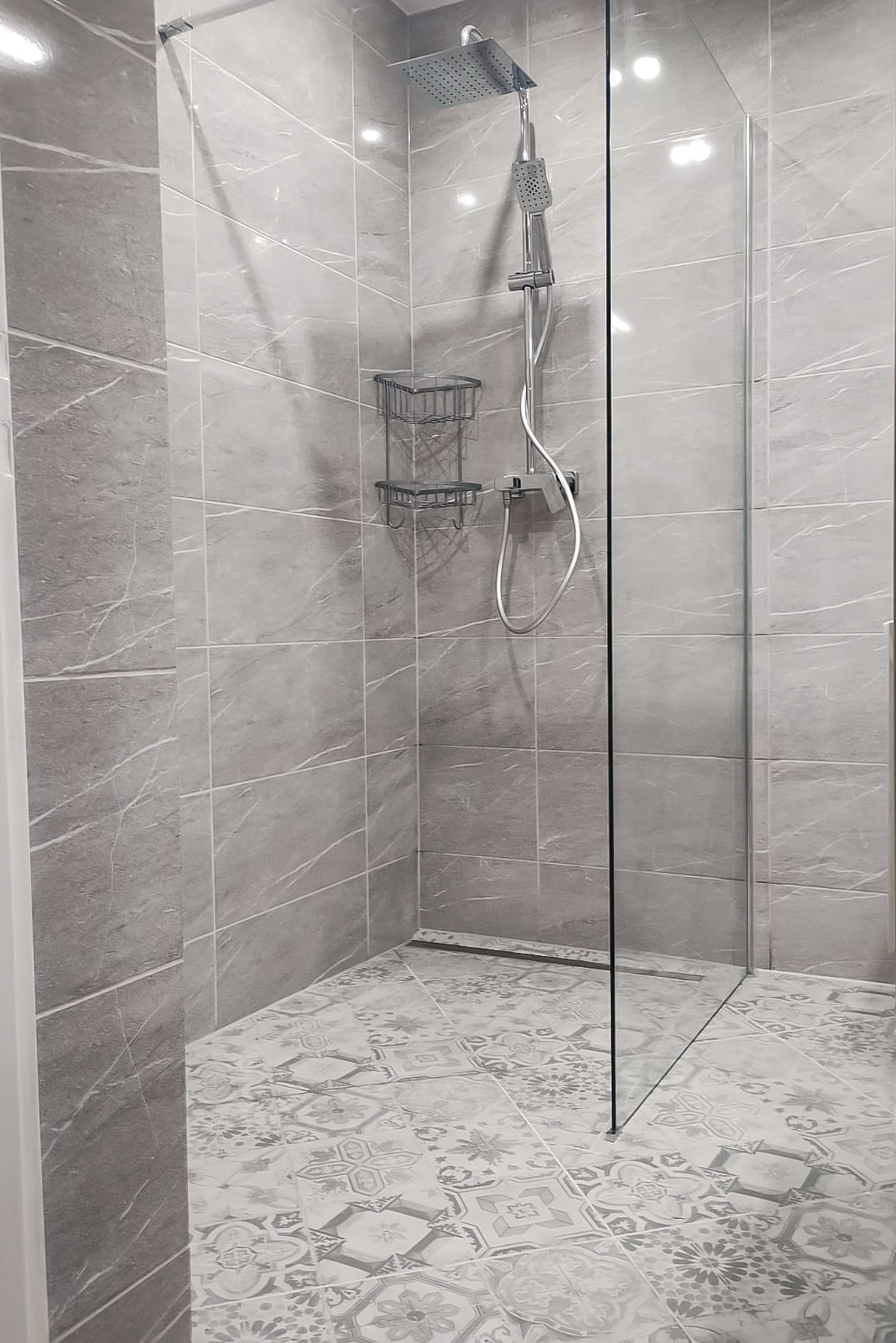 Nowoczesna kabina prysznicowa walk-in z szarymi płytkami imitującymi marmur i wzorzystą podłogą, wyposażona w deszczownicę i półkę na kosmetyki.
