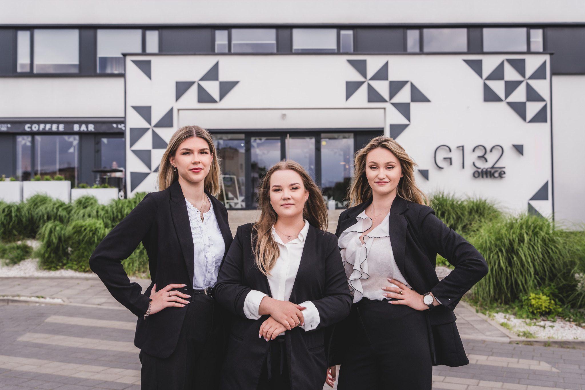 Trzy kobiety w garniturach stoją przed nowoczesnym biurowcem G132 Office z geometrycznym wzorem na elewacji i napisem 'Coffee Bar' w tle.