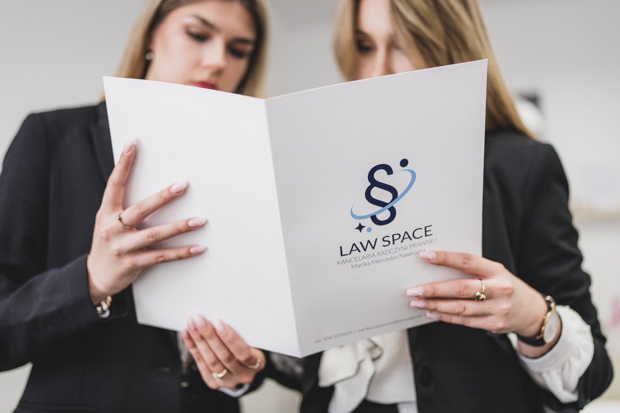 Dwie kobiety w garniturach przeglądają otwartą teczkę z logo kancelarii 'Law Space' w jasnym wnętrzu. Skupienie na dokumencie, profesjonalny wizerunek.