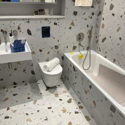 Art - Nowoczesna łazienka z terazzo na podłodze i ścianach, biała wanna z szarą matą antypoślizgową, wisząca toaleta z ręcznikiem, lustro z szafką i półką, widoczna instalacja elektryczna.