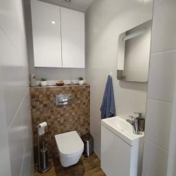 Art - Nowoczesna, mała łazienka z wiszącą toaletą, umywalką nablatową i szafką wiszącą. Ściana za toaletą wykończona mozaiką z drewnianych kostek, a pozostałe ściany białymi, błyszczącymi płytkami.