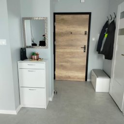 Art - Nowoczesny przedpokój z drewnopodobnymi drzwiami wejściowymi, szarą podłogą, białymi meblami, lustrem w srebrnej ramie i wieszakami na ścianie.