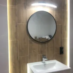 Nowoczesna umywalka z białą szafką, okrągłe lustro z czarną ramą podświetlone lampą LED, płytki imitujące drewno z oświetleniem LED po bokach w wyremontowanej łazience.