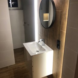 Art - Nowoczesna łazienka z umywalką na szafce, okrągłym lustrem z czarną ramą, oświetleniem LED i płytkami imitującymi drewno na podłodze i ścianie.