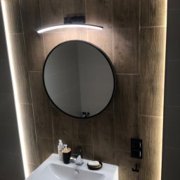 Art - Nowoczesna łazienka z okrągłym lustrem podświetlonym od góry, drewnopodobne płytki na ścianach z dekoracyjnym oświetleniem LED po bokach, biała umywalka z minimalistyczną armaturą i akcesoriami.