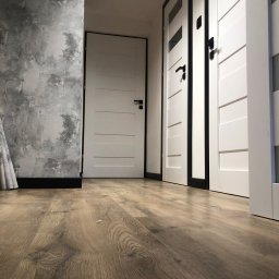 Art - Nowoczesne, białe drzwi z czarnymi detalami, widoczne z perspektywy podłogi z paneli w odcieniach brązu, ściana z efektem betonu w tle.