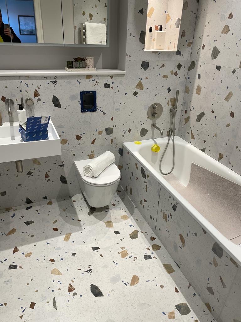 Nowoczesna łazienka z terazzo na podłodze i ścianach, biała wanna z szarą matą antypoślizgową, wisząca toaleta z ręcznikiem, lustro z szafką i półką, widoczna instalacja elektryczna.