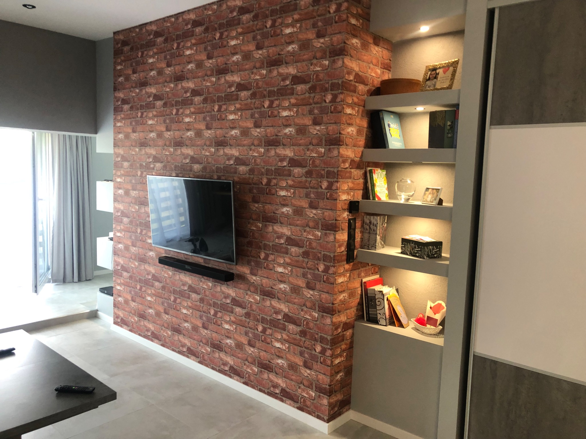 Ściana akcentowa z cegły klinkierowej w salonie z telewizorem i wnęką z półkami podświetlanymi LED, dekoracje i książki na półkach.