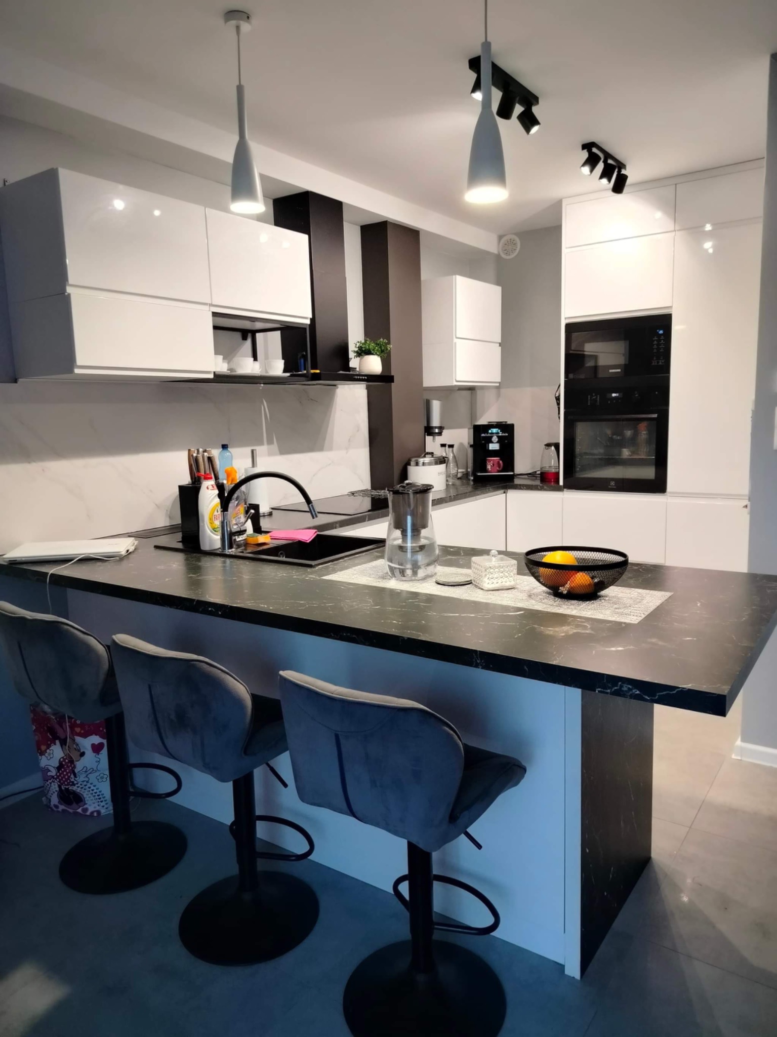 Nowoczesna kuchnia z wyspą, białe szafki z połyskiem, czarny blat imitujący marmur, trzy szare hokery, oświetlenie punktowe i wiszące lampy.