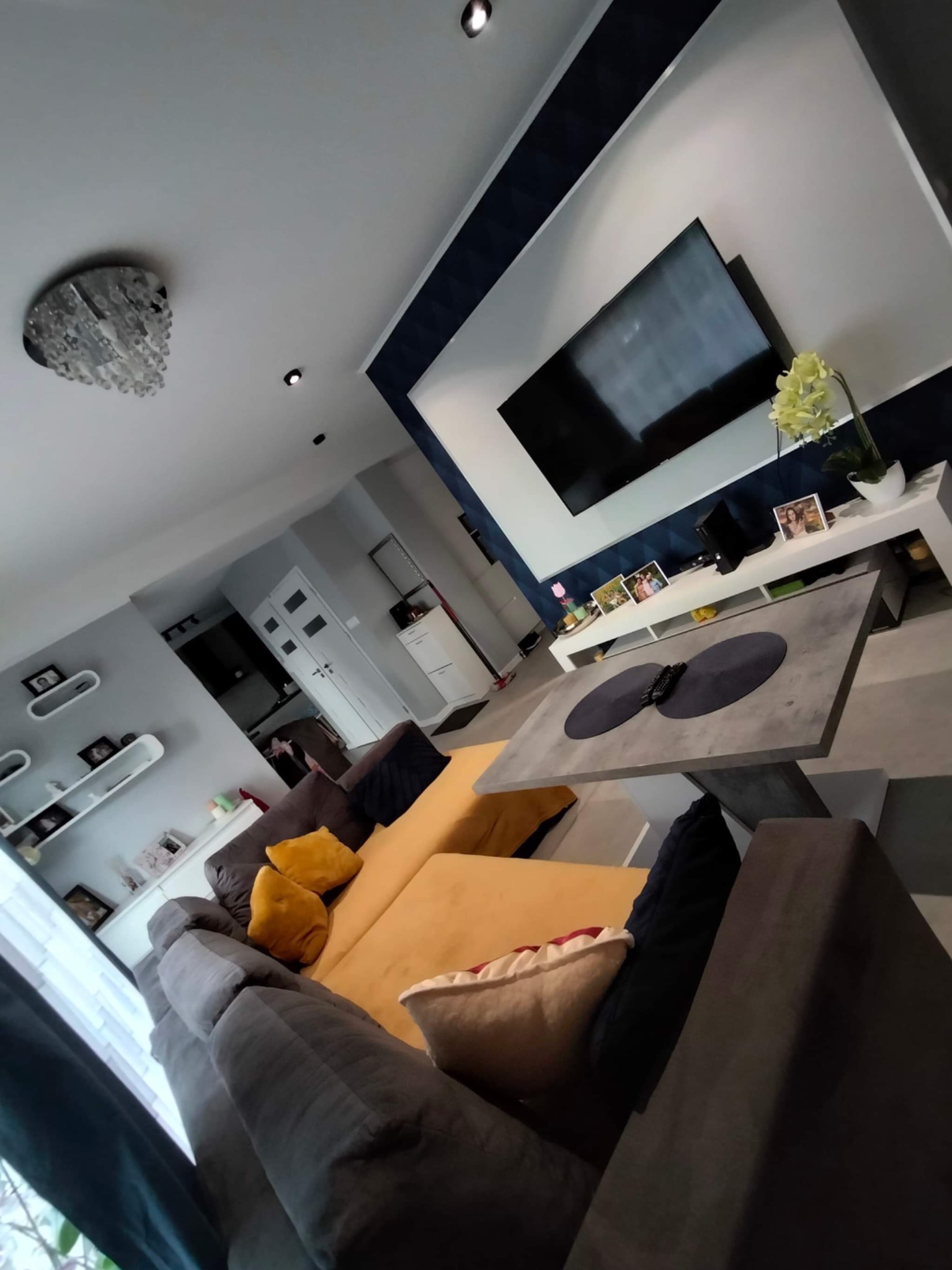 Nowoczesny salon z szarym narożnikiem, żółtymi poduszkami, betonowym stolikiem kawowym i telewizorem na ścianie z dekoracyjnym pasem.