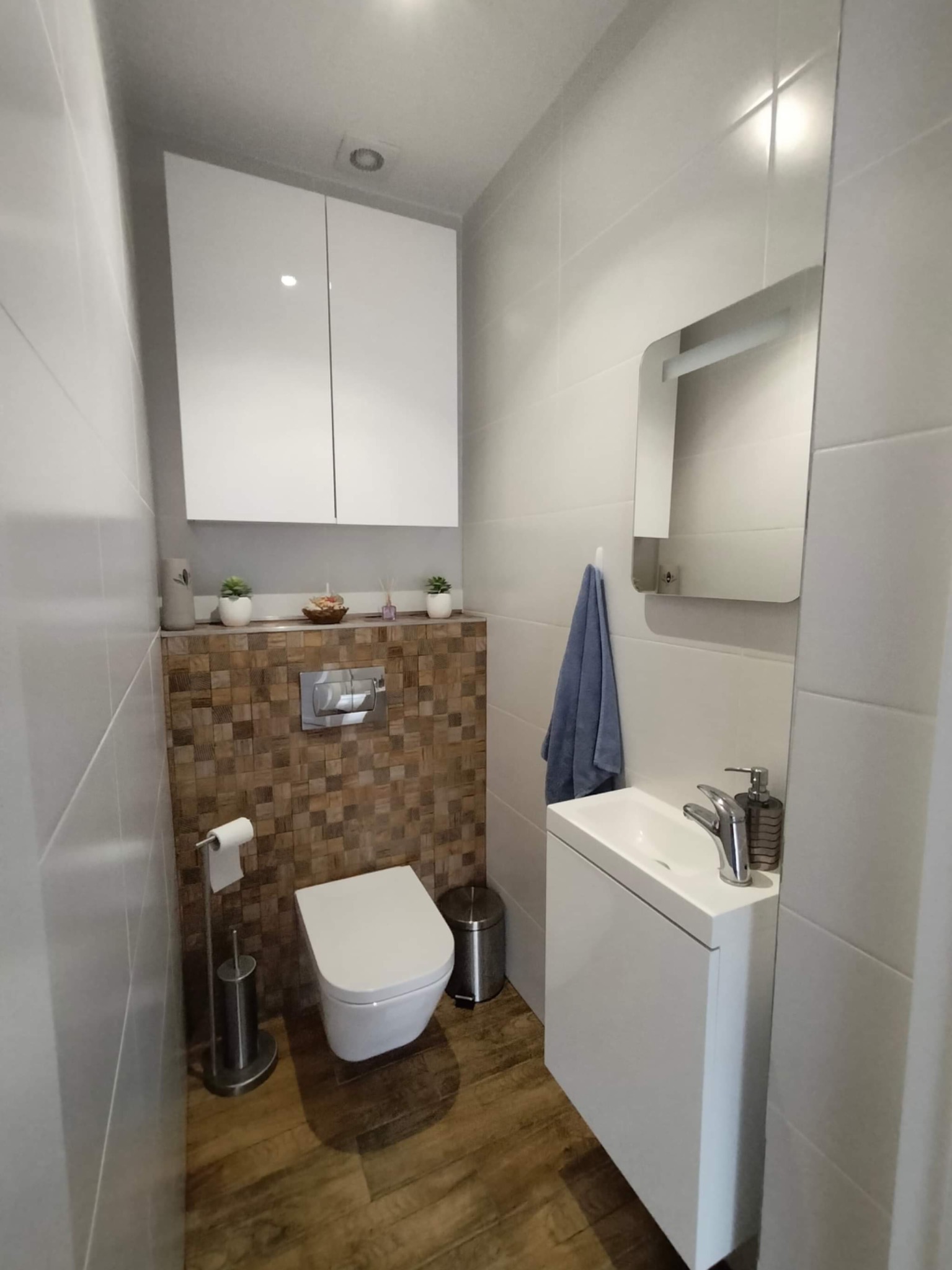 Nowoczesna, mała łazienka z wiszącą toaletą, umywalką nablatową i szafką wiszącą. Ściana za toaletą wykończona mozaiką z drewnianych kostek, a pozostałe ściany białymi, błyszczącymi płytkami.