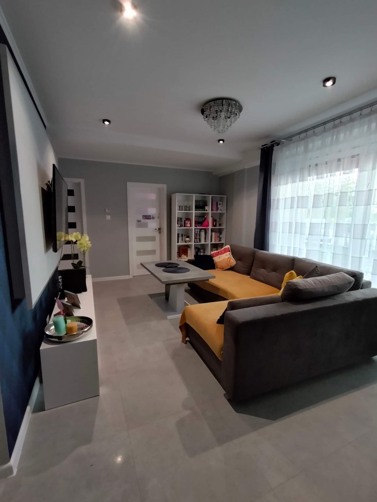 Nowoczesny salon z narożną sofą w odcieniach szarości i żółci, podwieszanym kryształowym żyrandolem i ścianami w dwóch odcieniach szarości oddzielonych ciemnoniebieskim pasem.