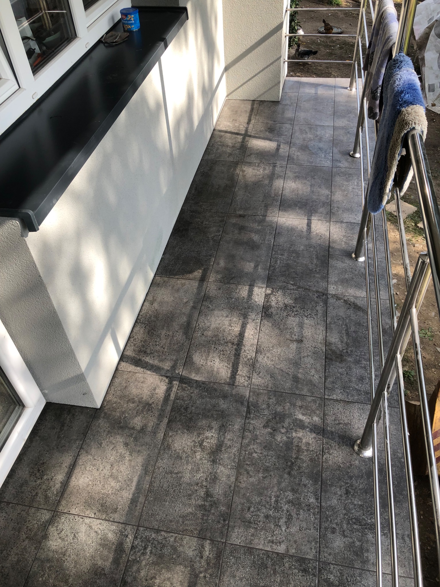 Wyłożony ciemnoszarą terakotą balkon z metalową balustradą, na której wiszą ręczniki, z widokiem na podwórze z kaczkami.