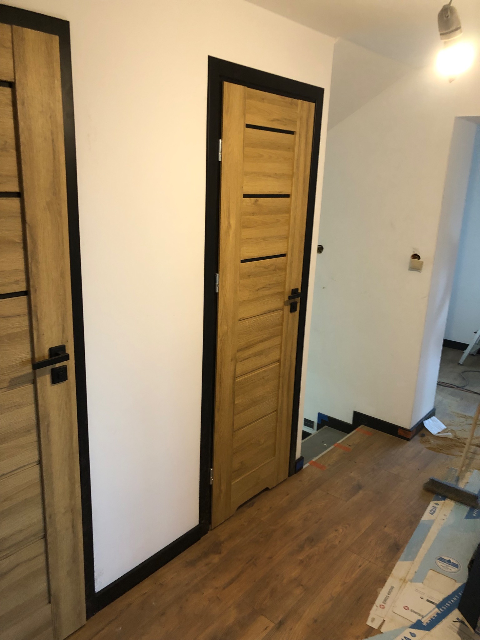 Dwa drewniane drzwi z czarną ramą i poziomymi panelami, zamontowane w białej ścianie, widoczne fragmenty podłogi i schodów w trakcie prac wykończeniowych.