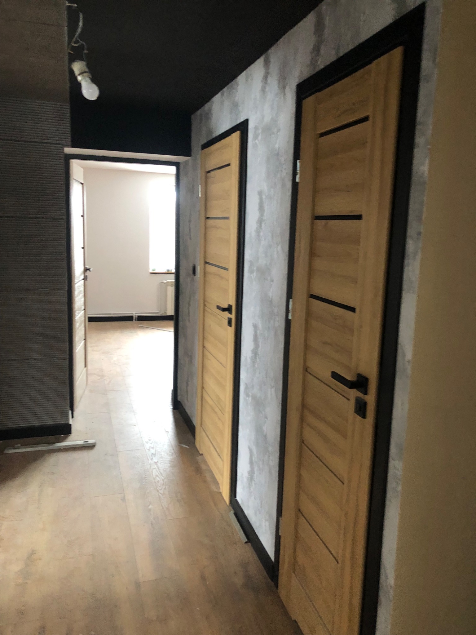 Dwa nowocześnie wykończone, jasne drewniane drzwi z czarnymi ramami i poziomymi wstawkami, zamontowane w szarej ścianie z efektem betonu, widoczne z perspektywy korytarza z drewnianą podłogą...