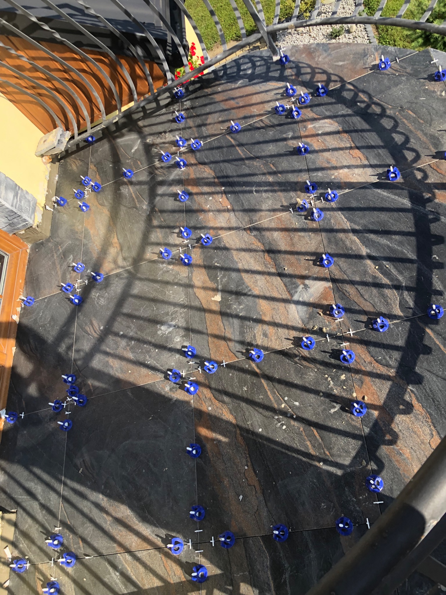 Układanie ciemnoszarych płytek na balkonie z użyciem systemu poziomowania, widoczne niebieskie klipsy i sznurki wyznaczające linie.