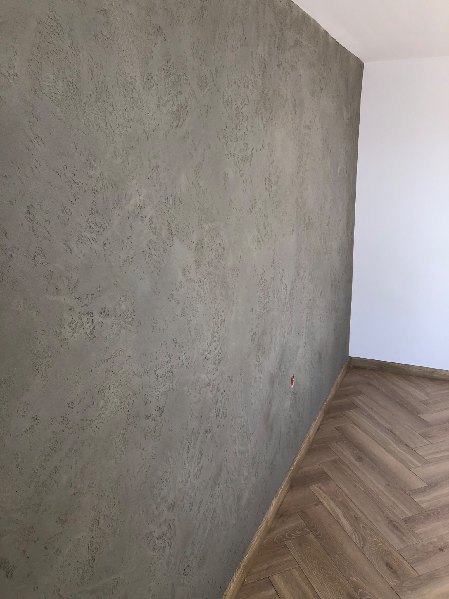 Szara ściana z dekoracyjnym tynkiem o nieregularnej fakturze, widoczna w rogu pomieszczenia z parkietem ułożonym w jodełkę i białą ścianą.