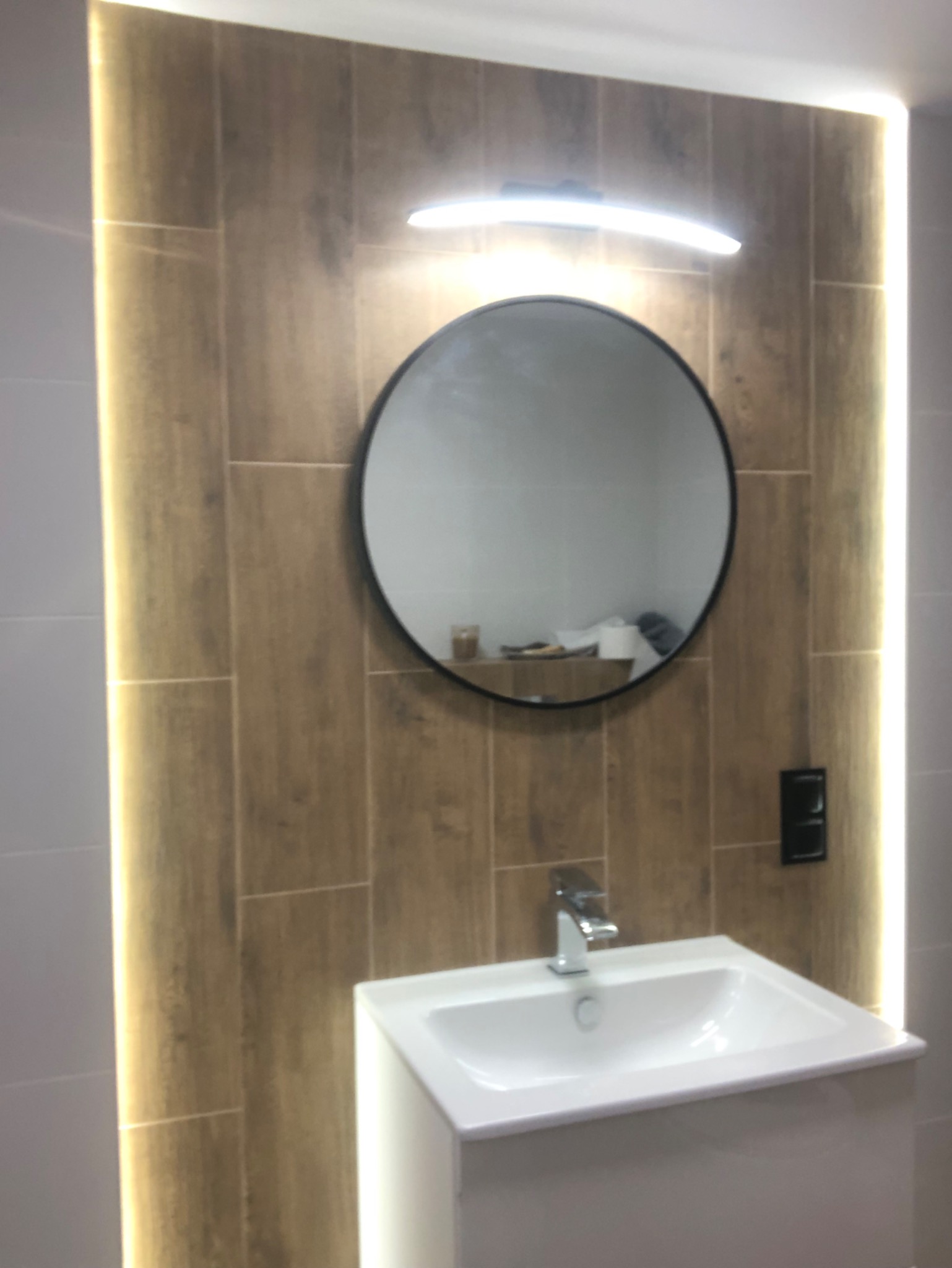 Nowoczesna umywalka z białą szafką, okrągłe lustro z czarną ramą podświetlone lampą LED, płytki imitujące drewno z oświetleniem LED po bokach w wyremontowanej łazience.
