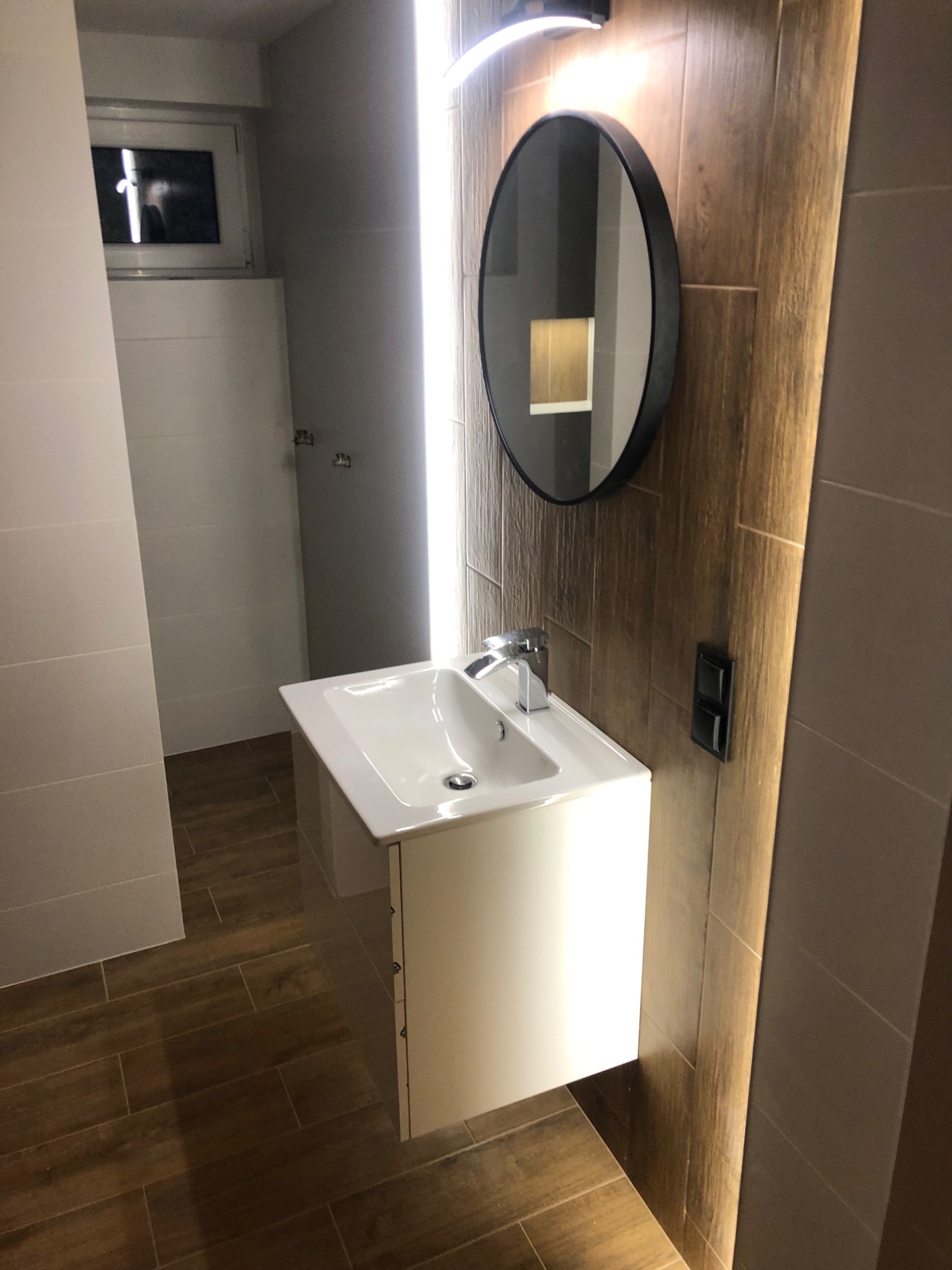 Nowoczesna łazienka z umywalką na szafce, okrągłym lustrem z czarną ramą, oświetleniem LED i płytkami imitującymi drewno na podłodze i ścianie.