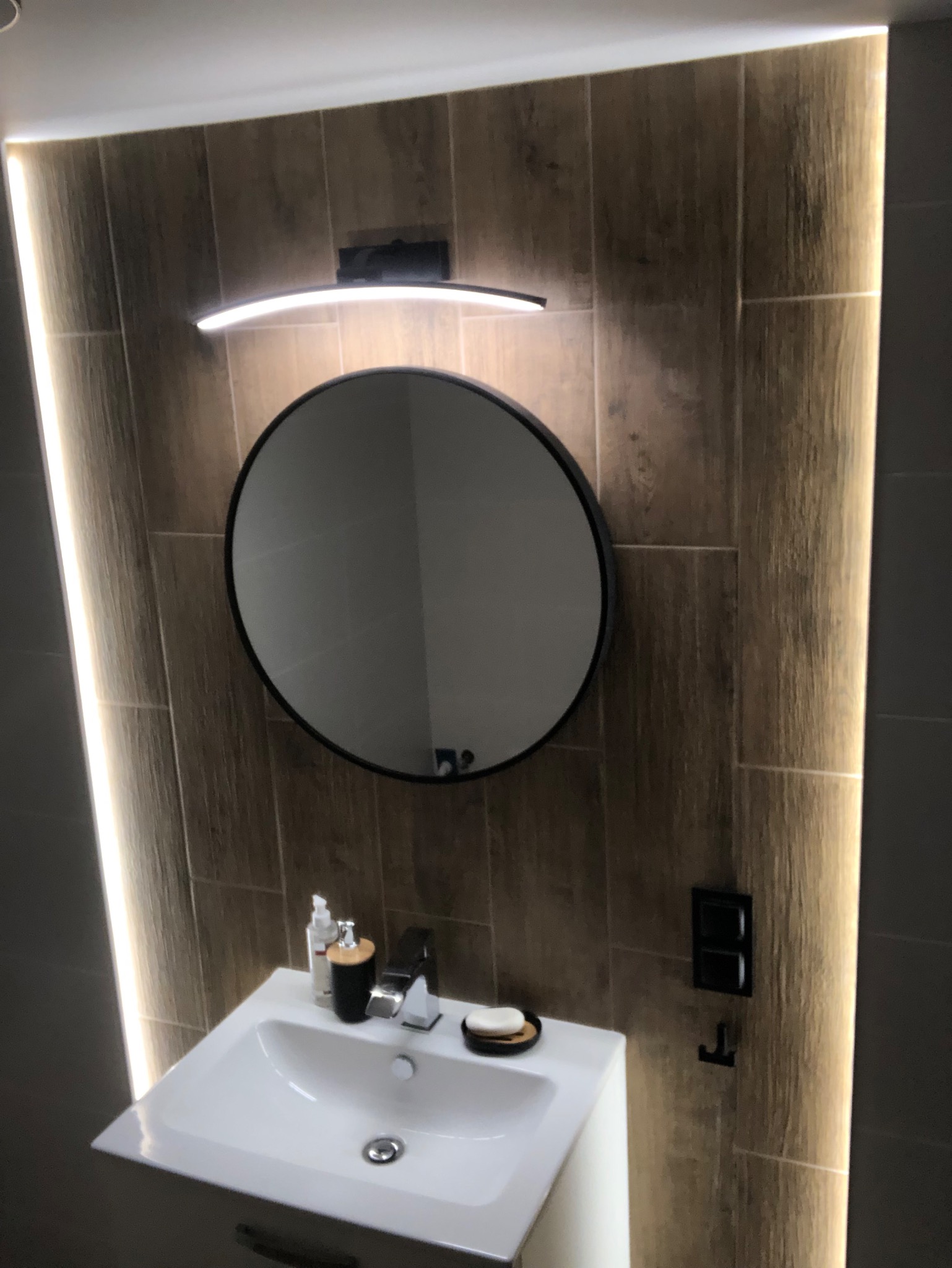Nowoczesna łazienka z okrągłym lustrem podświetlonym od góry, drewnopodobne płytki na ścianach z dekoracyjnym oświetleniem LED po bokach, biała umywalka z minimalistyczną armaturą i akcesoriami.