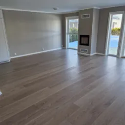 Jasne, przestronne wnętrze po remoncie z nową podłogą z paneli w odcieniach szarości, ścianami w kolorze taupe, białymi listwami przypodłogowymi i minimalistycznym kominkiem w narożniku.