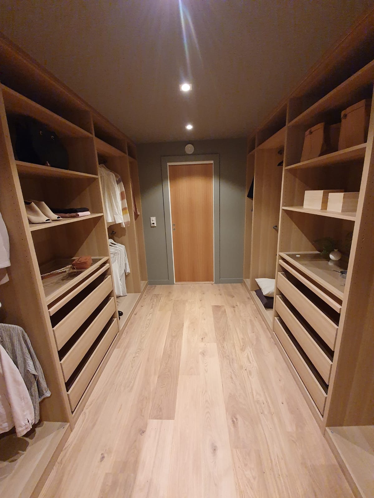 Wnętrze garderoby z drewnianymi szafami z szufladami i półkami, podłoga z jasnego drewna, drzwi w tle, oświetlenie sufitowe punktowe.