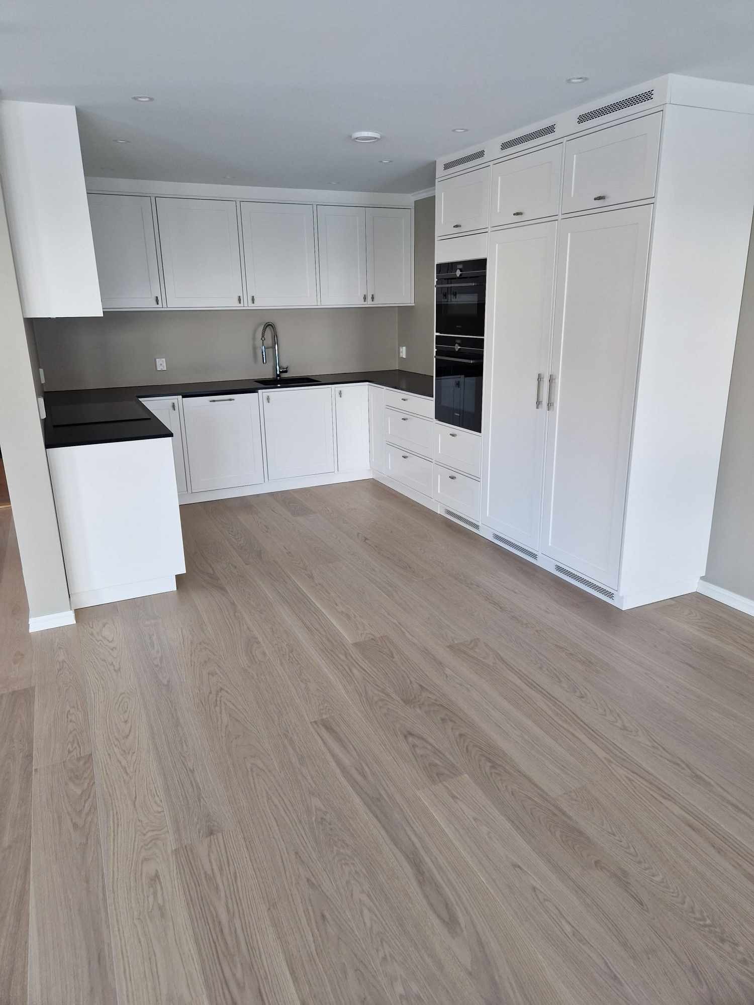 Jasna, minimalistyczna kuchnia z białymi szafkami i czarnym blatem, drewniana podłoga, zabudowane piekarniki.