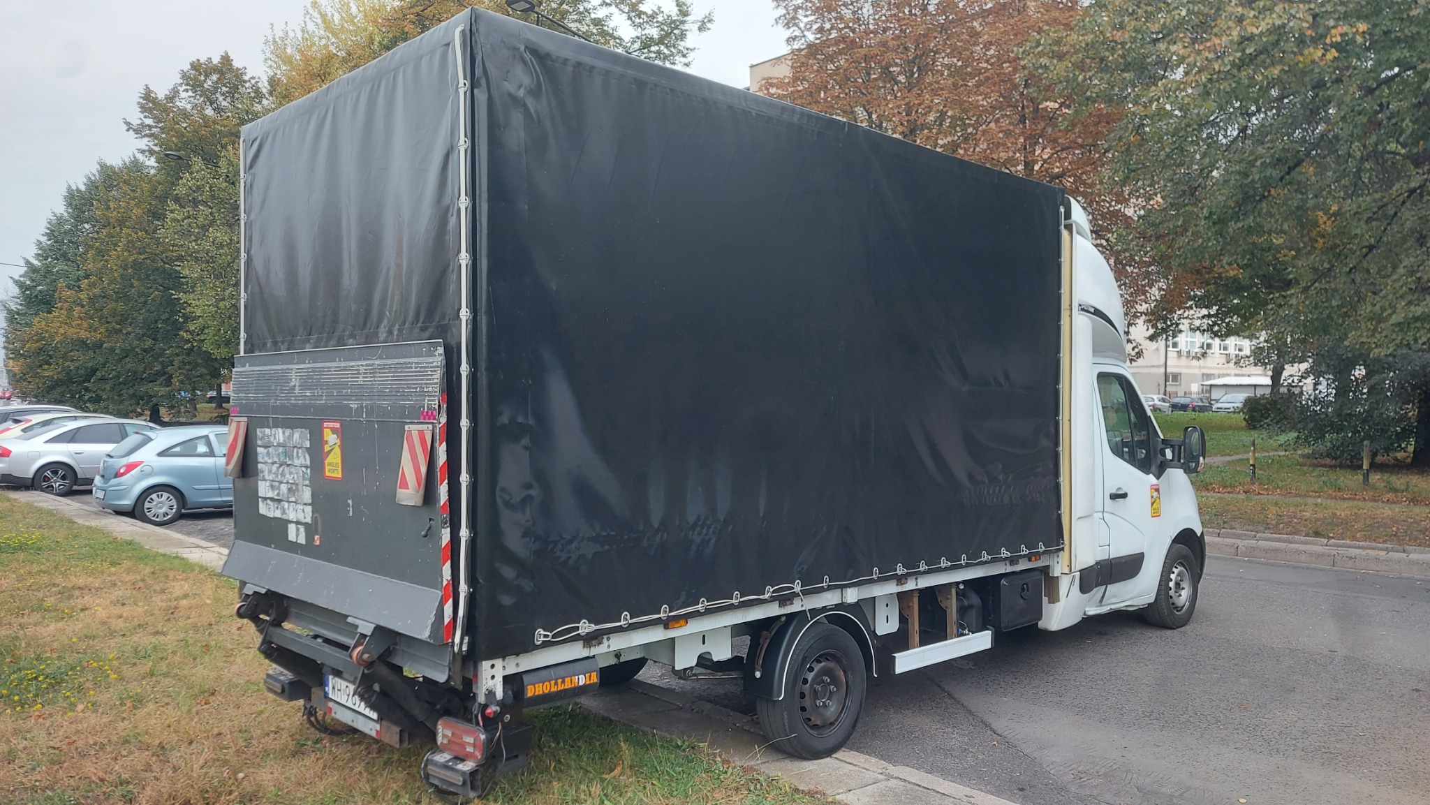 Biały samochód dostawczy z czarną plandeką, zaparkowany na poboczu w jesienny dzień, widoczne tablice ostrzegawcze na tylnej klapie.