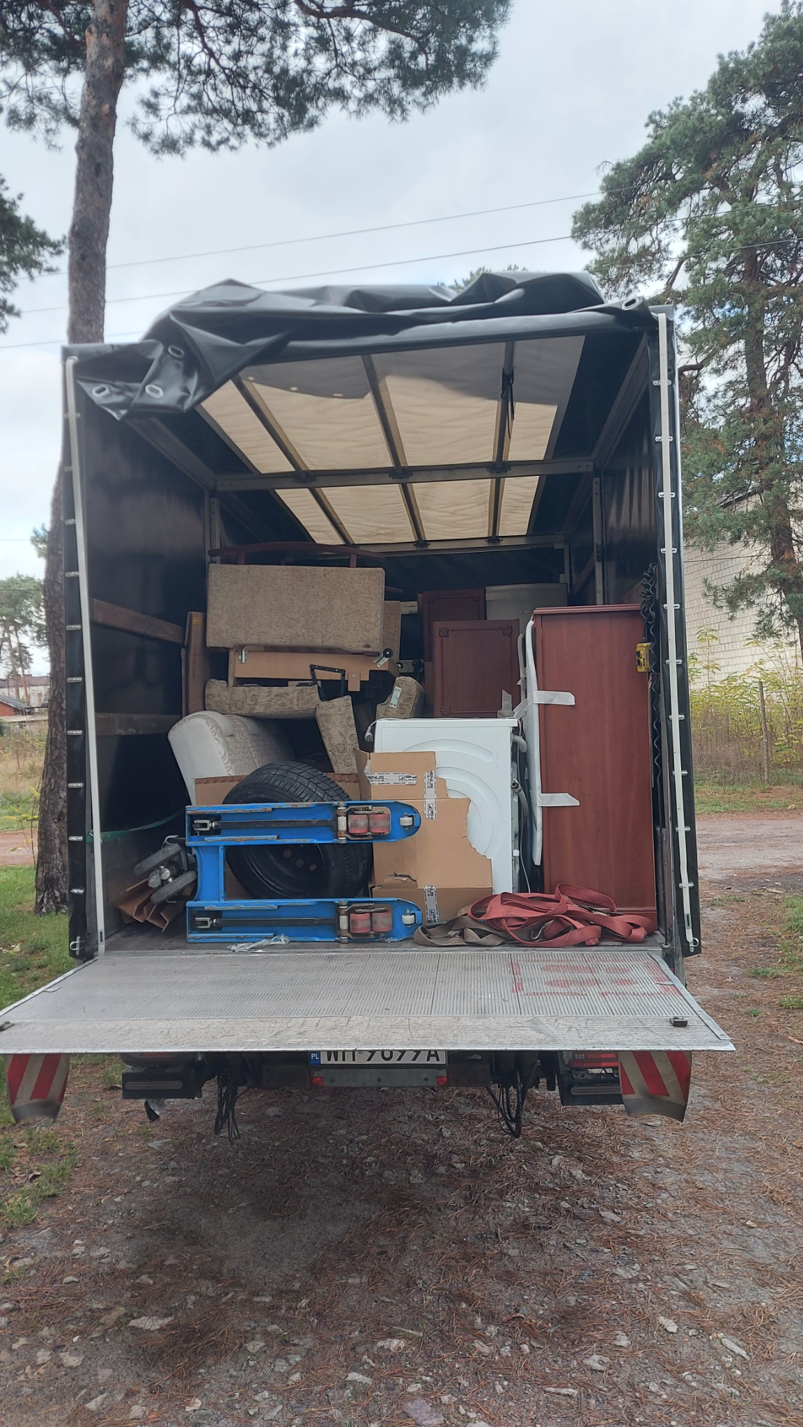 Otwarta paka samochodu dostawczego załadowana meblami, sprzętem AGD i kartonami, zabezpieczona czarną plandeką, widoczne niebieskie wózki transportowe i tablica rejestracyjna pojazdu.