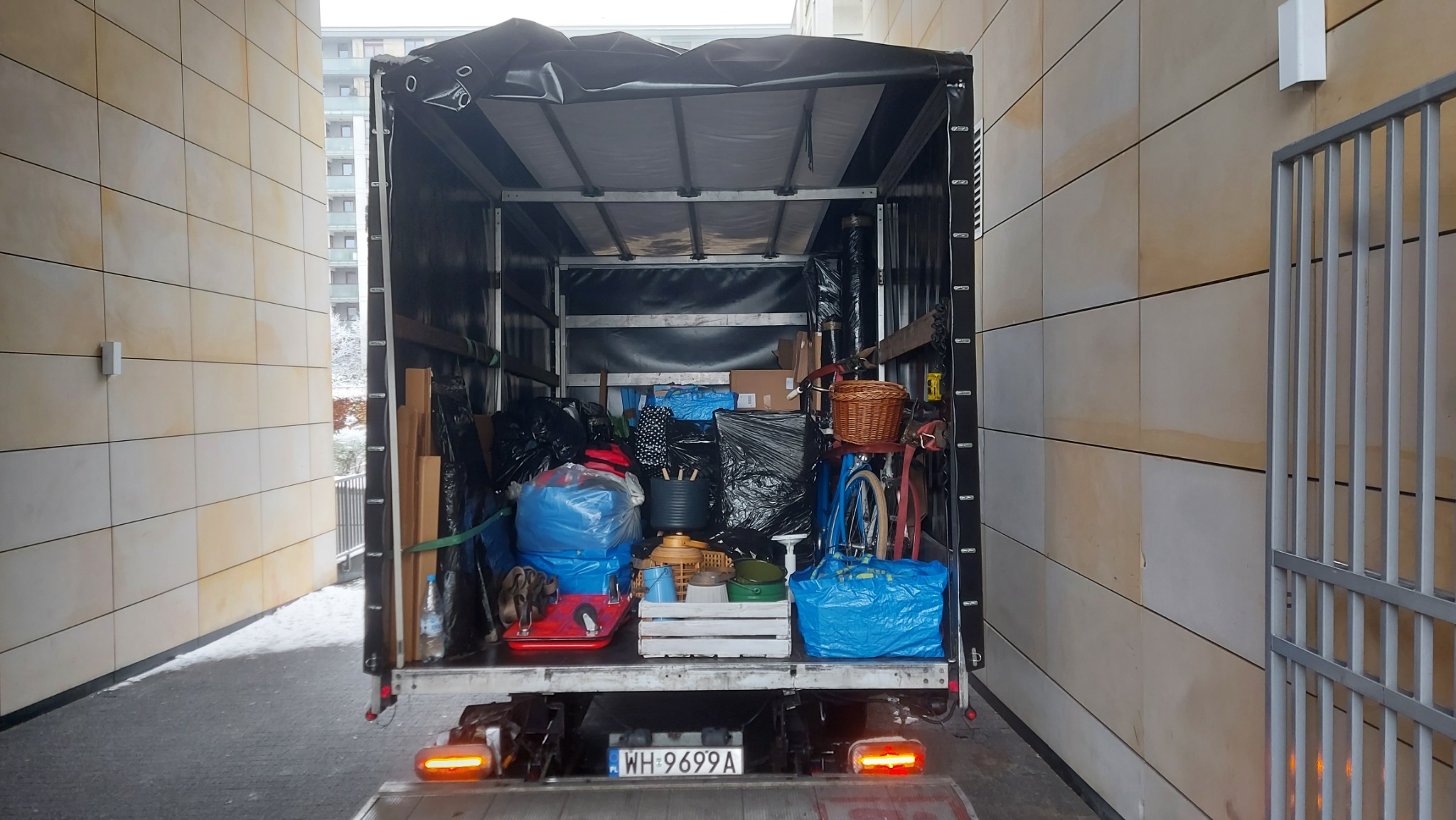 Wnętrze skrzyni ładunkowej wypełnionej różnorodnymi przedmiotami, w tym workami, pudełkami, koszami i rowerem, przygotowanymi do transportu podczas przeprowadzki. Widoczne tablice rejestracyjne...