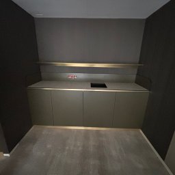 MULTITASK Sergiusz Kostenski - Minimalistyczna zabudowa szafkowa w ciemnej tonacji z blatem i złotymi akcentami, widoczna instalacja elektryczna nad blatem, surowe wnętrze bez dekoracji.