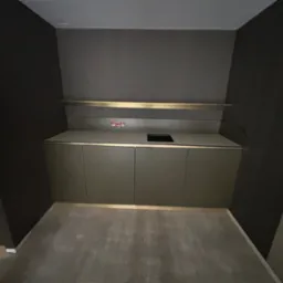 Minimalistyczna zabudowa szafkowa w ciemnej tonacji z blatem i złotymi akcentami, widoczna instalacja elektryczna nad blatem, surowe wnętrze bez dekoracji.