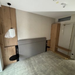 MULTITASK Sergiusz Kostenski - Pokój hotelowy w trakcie wykańczania: drewniana boazeria za łóżkiem z szarą tapicerką, wisząca lampa z plastikową osłoną, szafa i wieszak na ubrania z półką, jasna wykładzina dywanowa.