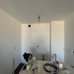 MULTITASK Sergiusz Kostenski - Wnętrze kuchni w trakcie remontu, świeżo pomalowane ściany na biało, widoczne puszki elektryczne, niepodłączone oświetlenie sufitowe, zlew z akcesoriami i środkami czystości na blacie...