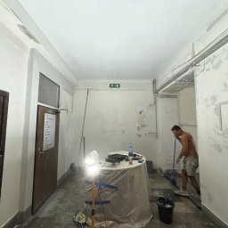 MULTITASK Sergiusz Kostenski - Mężczyzna bez koszulki, w szortach, myje podłogę w korytarzu, gdzie ściany są częściowo zagruntowane i przygotowane do malowania, widoczny stół z narzędziami malarskimi i oświetleniem halogenowym.