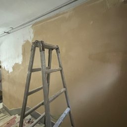 MULTITASK Sergiusz Kostenski - Ściana w trakcie malowania, widoczna aluminiowa drabina, fragment pomalowanej na biało powierzchni i rury pod sufitem.