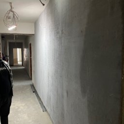 MULTITASK Sergiusz Kostenski - Ściana w trakcie szpachlowania z widoczną siatką wzmacniającą, surowy beton na podłodze, widok w głąb korytarza z otwartymi drzwiami, prowizoryczne oświetlenie zwisające z sufitu.