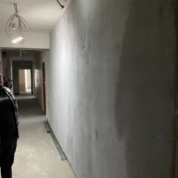 Ściana w trakcie szpachlowania z widoczną siatką wzmacniającą, surowy beton na podłodze, widok w głąb korytarza z otwartymi drzwiami, prowizoryczne oświetlenie zwisające z sufitu.