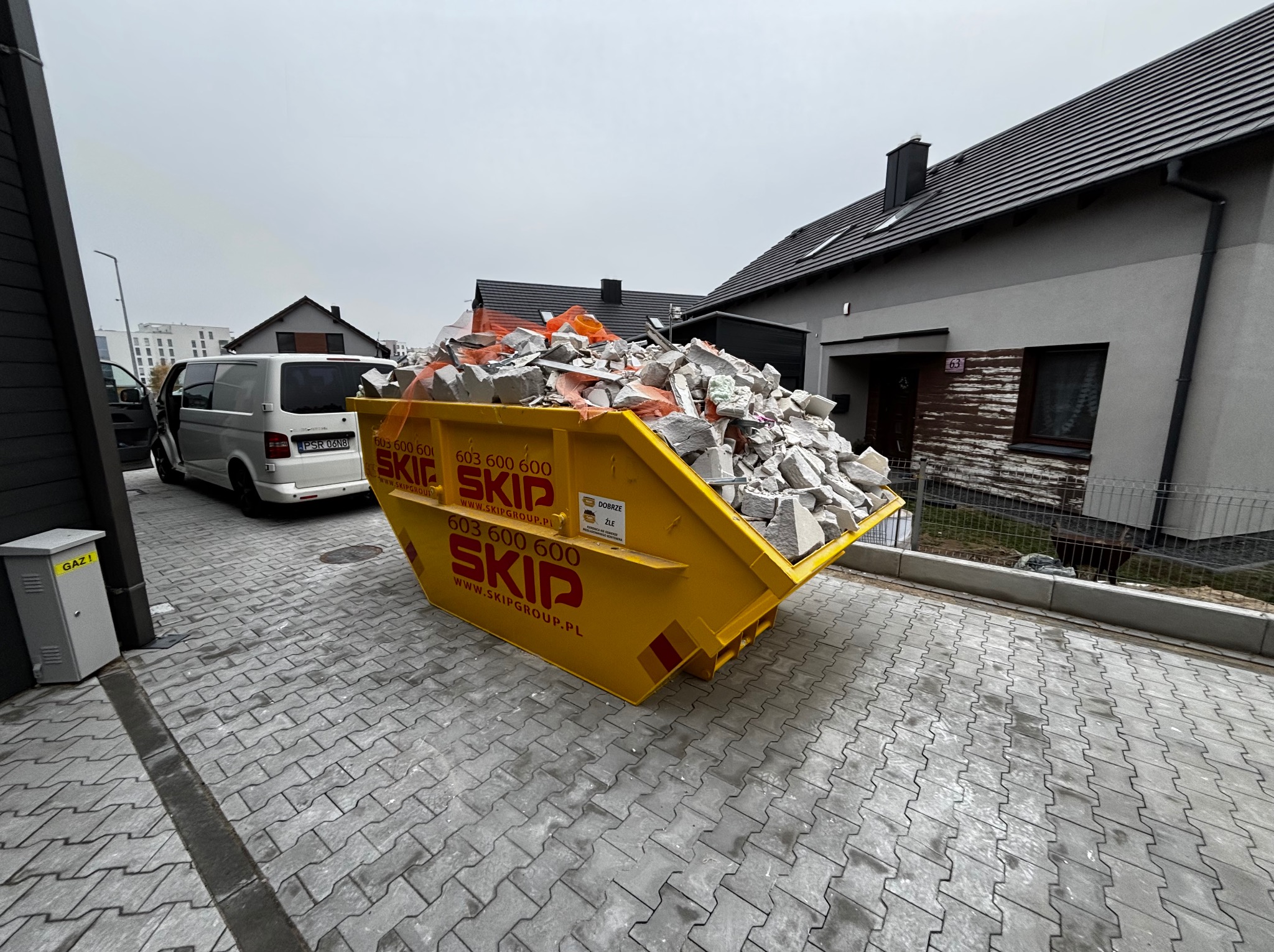 Żółty kontener SKIP wypełniony gruzem budowlanym stoi na szarej kostce brukowej przed domem jednorodzinnym z szarą elewacją i białym vanem z otwartymi drzwiami w tle.