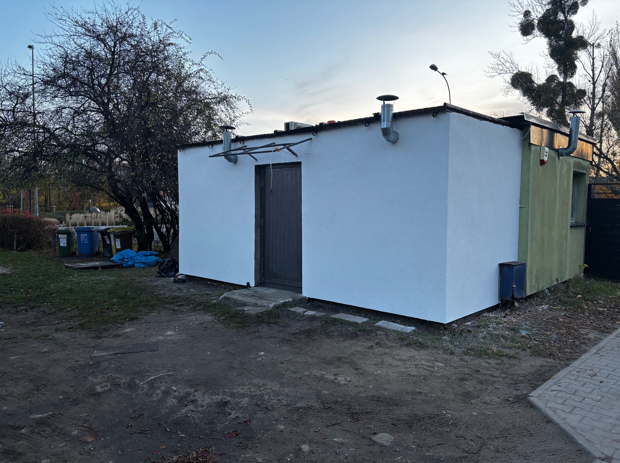 Otynkowany, parterowy budynek z dwoma kominami i ciemnymi drzwiami wejściowymi, widoczne ślady po niedawnym malowaniu elewacji.