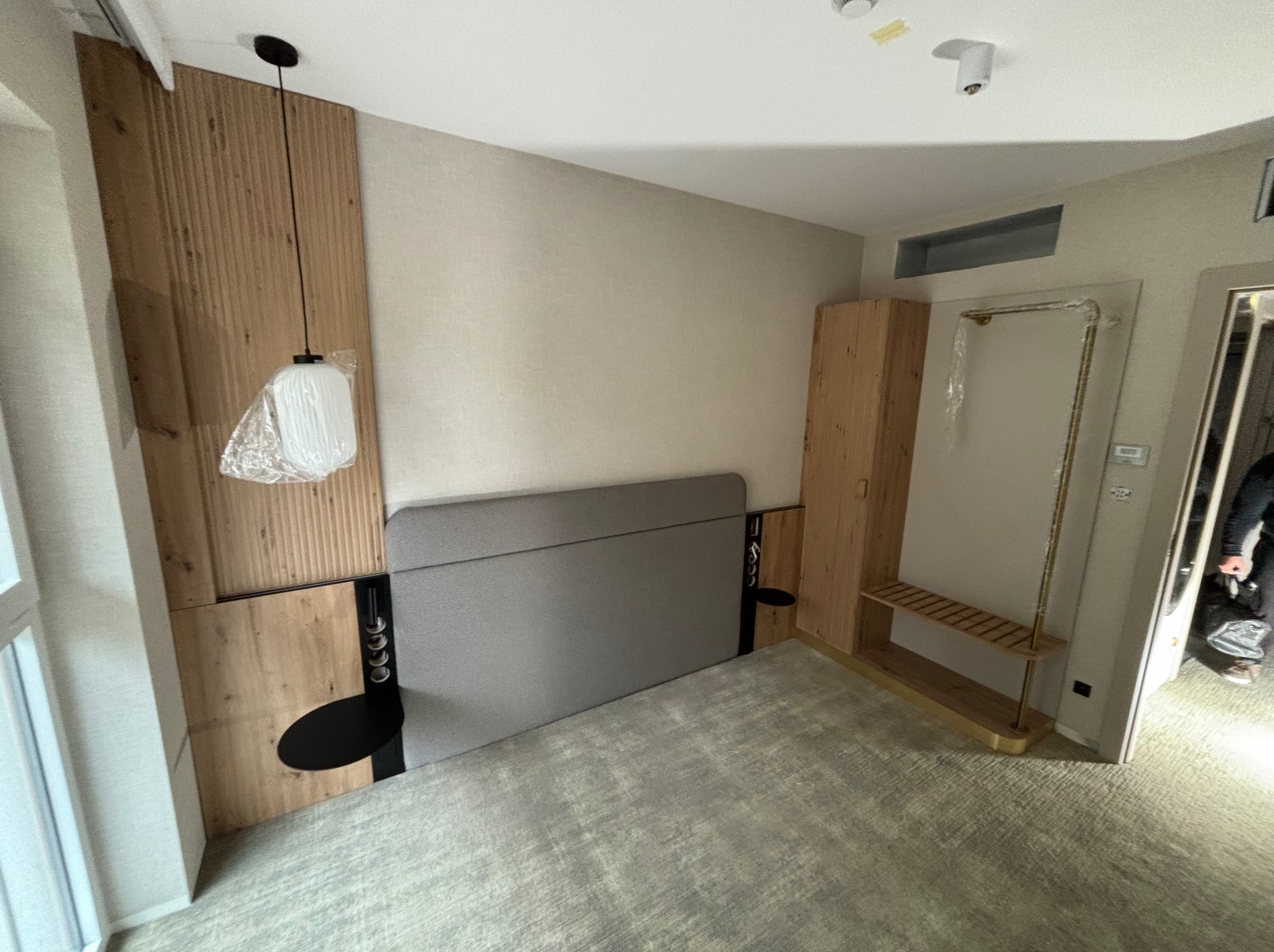 Pokój hotelowy w trakcie wykańczania: drewniana boazeria za łóżkiem z szarą tapicerką, wisząca lampa z plastikową osłoną, szafa i wieszak na ubrania z półką, jasna wykładzina dywanowa.