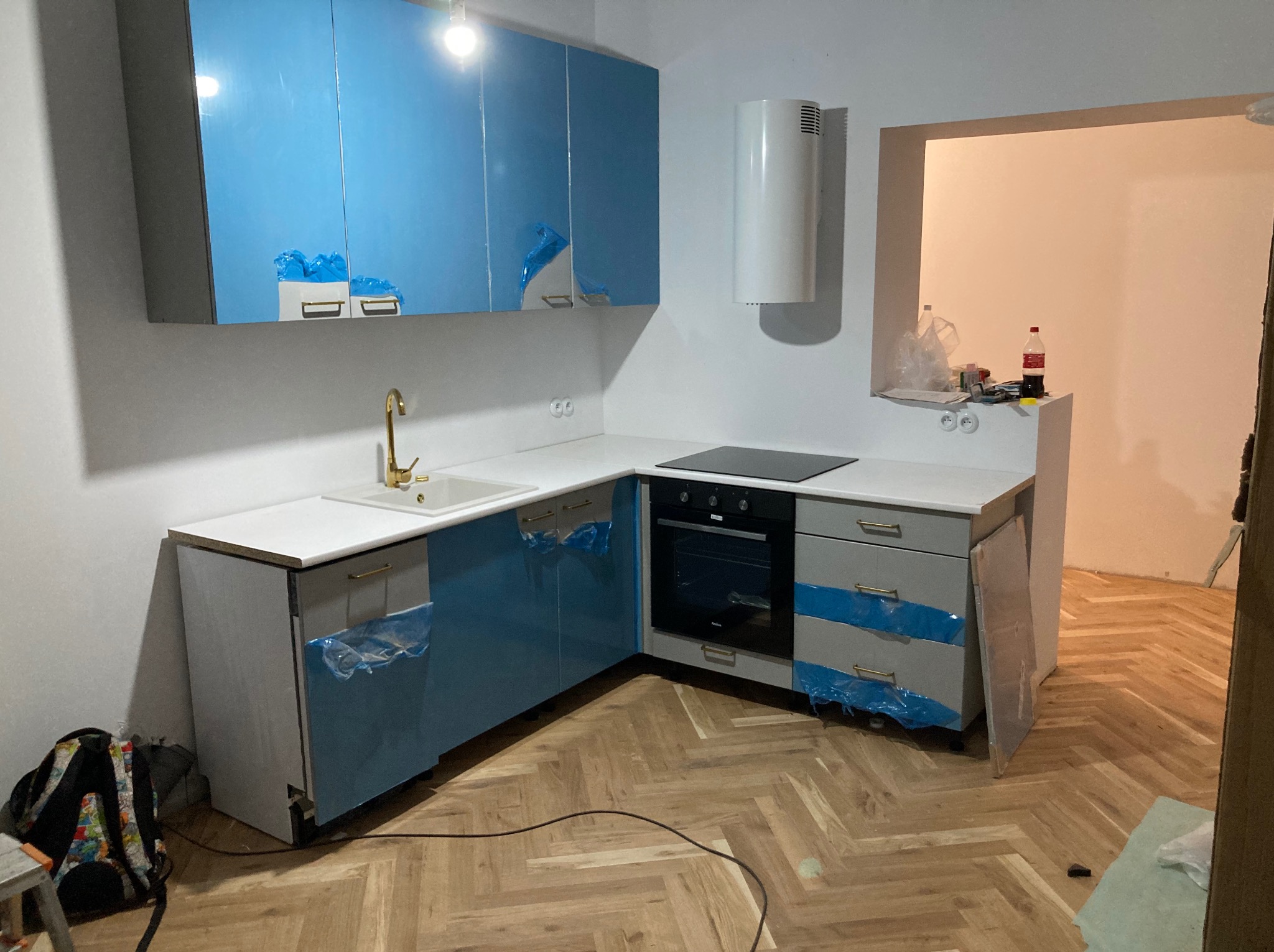 Nowoczesna, niebiesko-szara zabudowa kuchenna z białym blatem, jeszcze zabezpieczona folią ochronną, złota bateria umywalkowa, widok na wnętrze remontowanego mieszkania w Poznaniu.