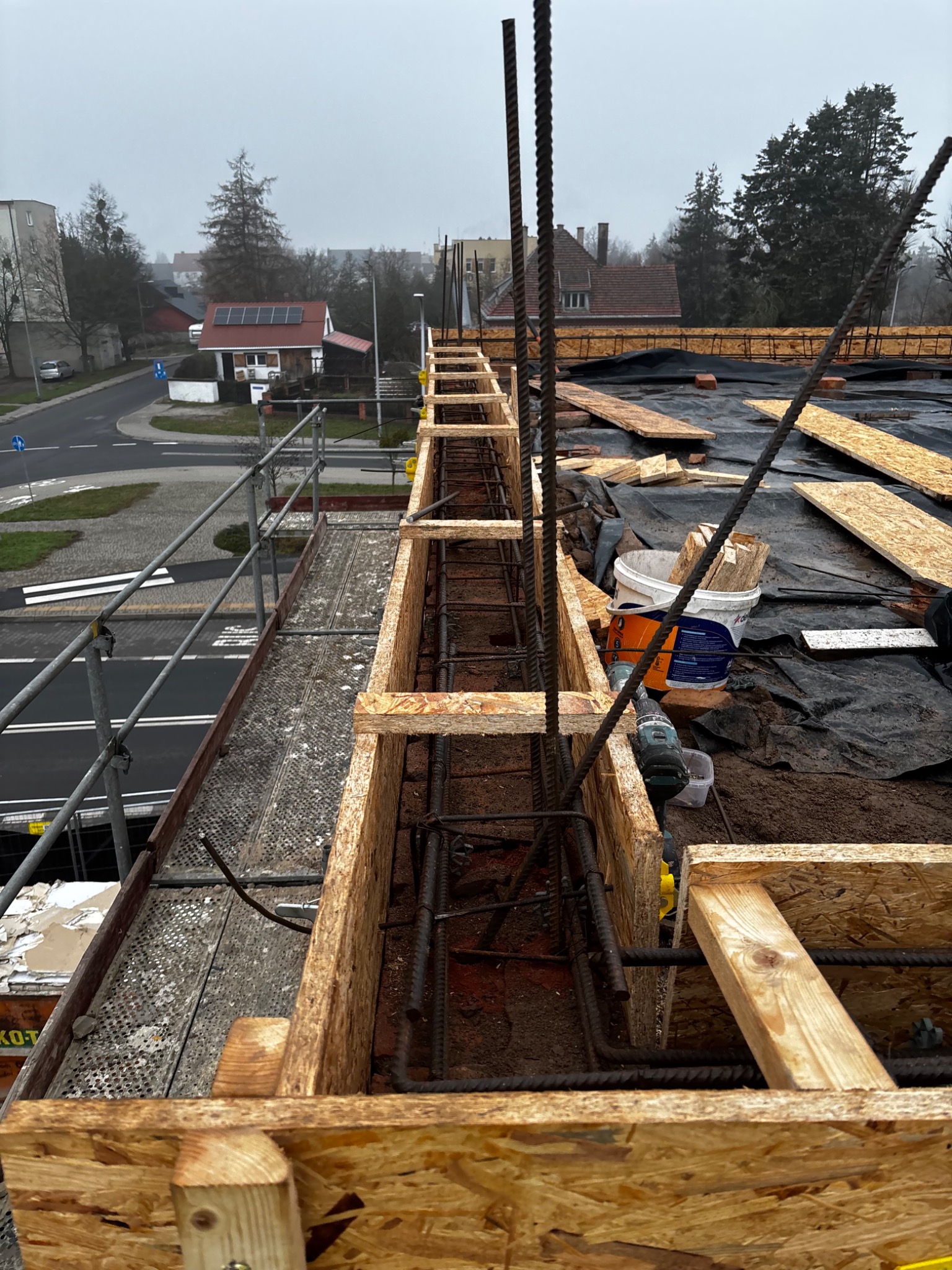Szczegółowe ujęcie szalunku zbrojeniowego na budowie, widoczne pręty zbrojeniowe, deski szalunkowe, narzędzia i wiadro z farbą, w tle zabudowa miejska w pochmurny dzień.