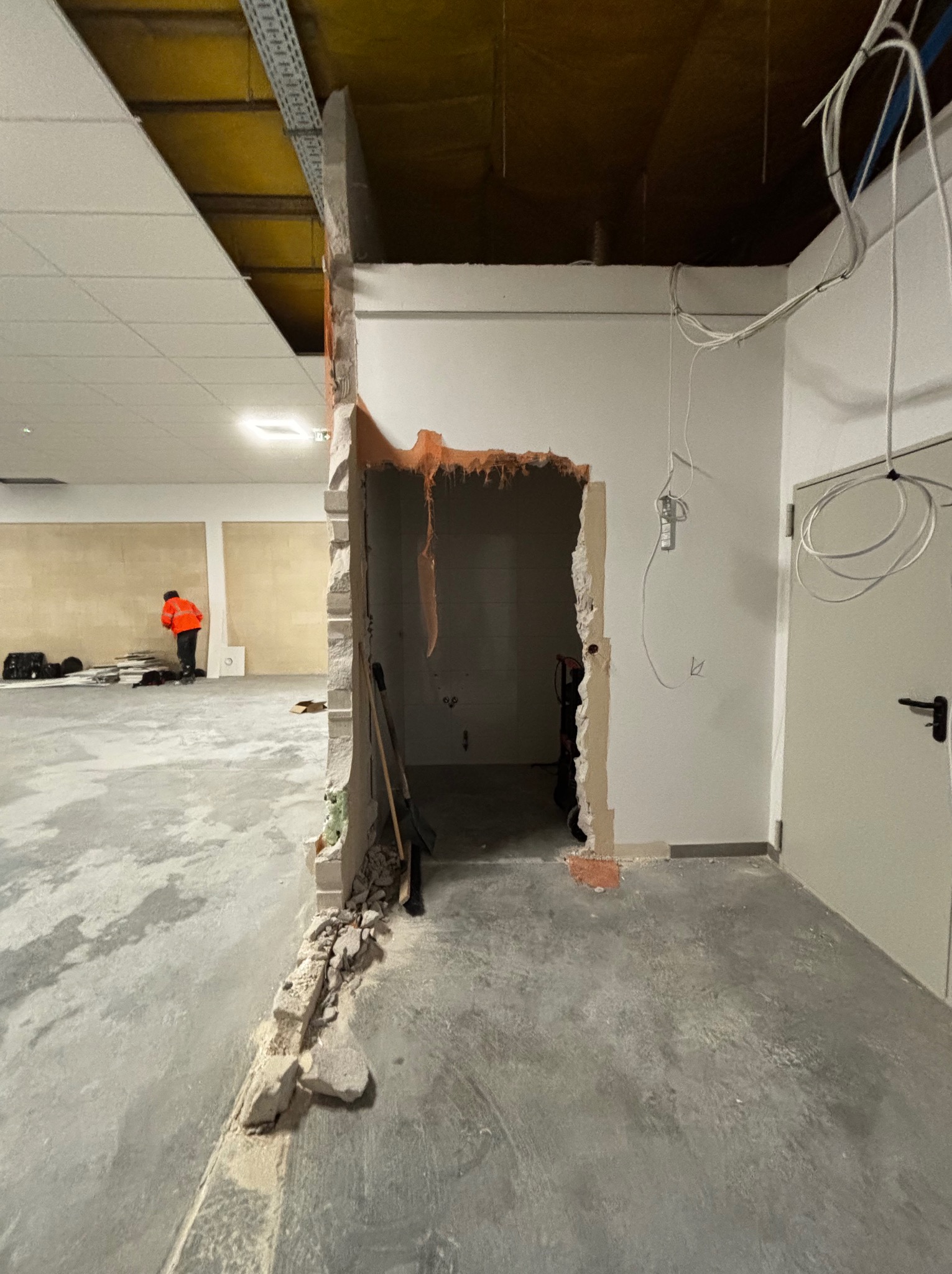 Wnętrze w trakcie prac rozbiórkowych: wyburzona ściana z widocznymi fragmentami gruzu na podłodze, wystające kable elektryczne, pracownik w pomarańczowej kurtce w tle.