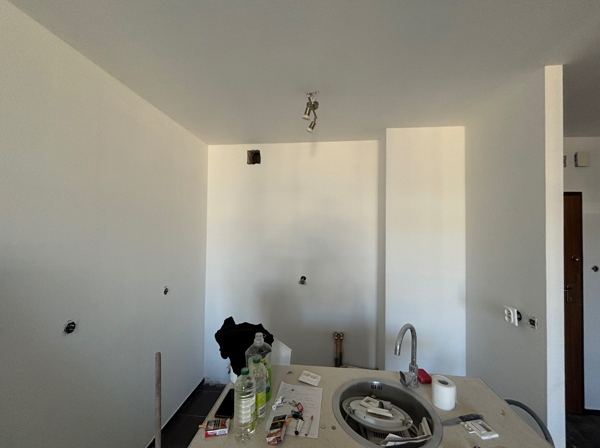 Wnętrze kuchni w trakcie remontu, świeżo pomalowane ściany na biało, widoczne puszki elektryczne, niepodłączone oświetlenie sufitowe, zlew z akcesoriami i środkami czystości na blacie...