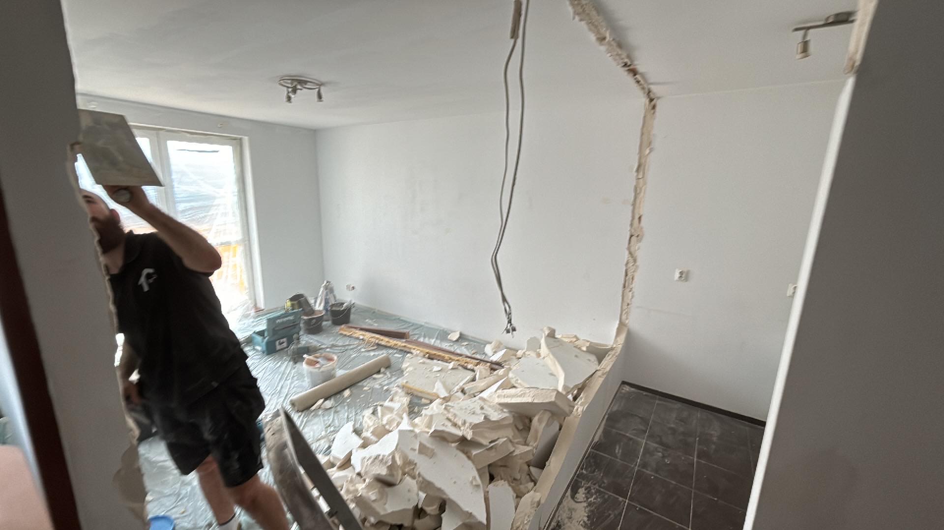 Pracownik z kielnią wyrównuje ścianę po wyburzeniu, widoczne gruzowisko i zabezpieczona folią podłoga w jasnym pomieszczeniu.