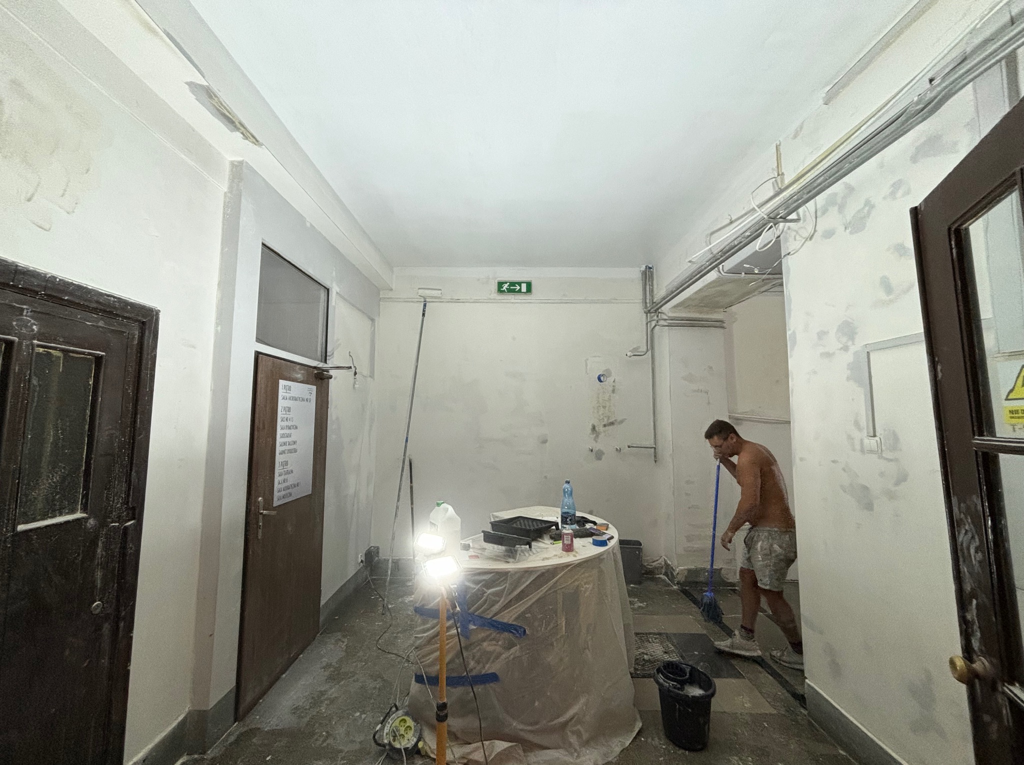 Mężczyzna bez koszulki, w szortach, myje podłogę w korytarzu, gdzie ściany są częściowo zagruntowane i przygotowane do malowania, widoczny stół z narzędziami malarskimi i oświetleniem halogenowym.