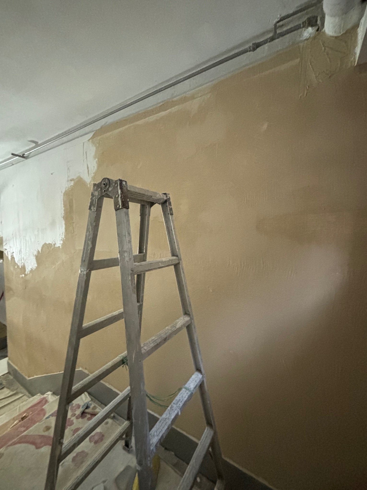 Ściana w trakcie malowania, widoczna aluminiowa drabina, fragment pomalowanej na biało powierzchni i rury pod sufitem.