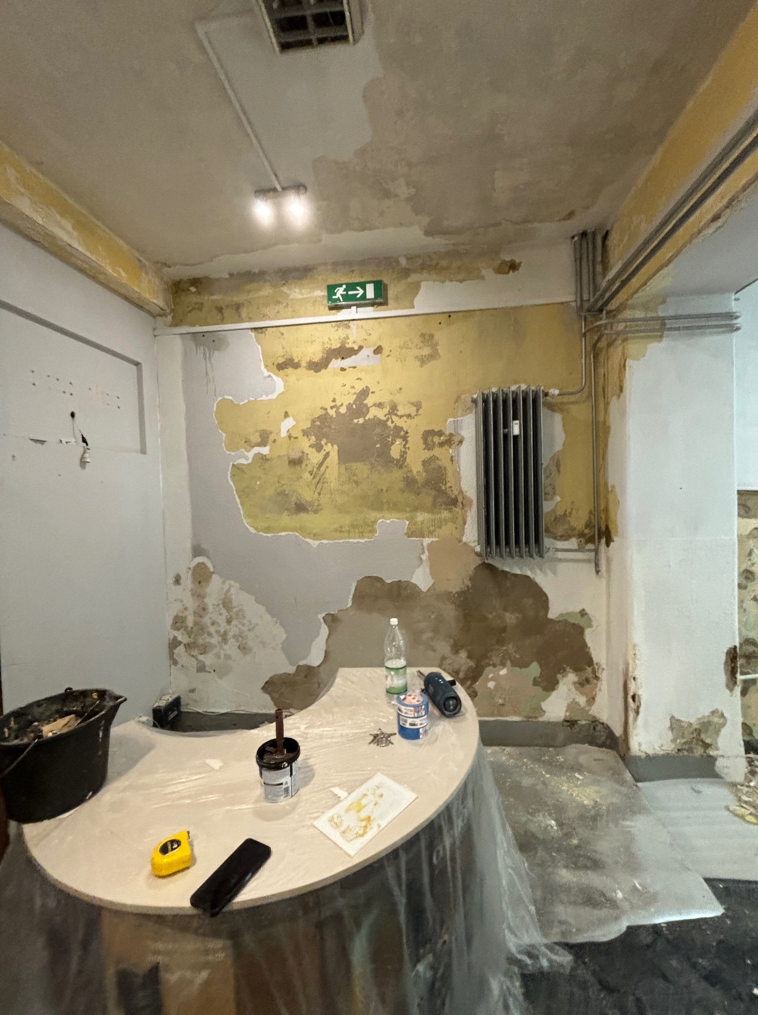 Przygotowanie do malowania pokoju: obskurne ściany z resztkami farby, grzejnik, stół z narzędziami malarskimi, wiadro z farbą, folia ochronna na podłodze, butelka wody i wiertarka.